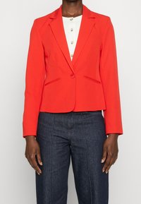 Blazer rouge sur mesure avec revers crantés, fermeture à un bouton et poches avant. Associé à un haut blanc et un pantalon en denim foncé.
