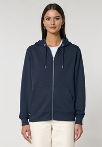 Felpa con zip di colore blu navy realizzata in tessuto morbido, con cordini, tasche frontali e polsini a costine. Indossata sopra una maglietta bianca e pantaloni color crema.