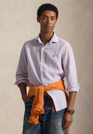 Camicia a righe viola chiaro con bottoni e maniche lunghe, abbinata a un maglione arancione lavorato a maglia annodato in vita e jeans di denim blu.