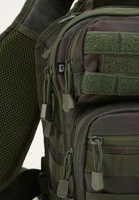 Camouflage backpack featuring textured fabric, multiple zippered pockets, green accents, and adjustable straps with mesh for breathability.