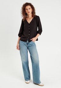 Schwarze, leichte Bluse mit V-Ausschnitt und Dreiviertelärmeln, verziert mit dekorativen Knöpfen auf der Vorderseite. Kombiniert mit blauen weit geschnittenen Jeans.