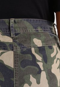 Bermudas de mezclilla con patrón de camuflaje en colores verde, marrón y marino. Presentan una cinturilla y bolsillos cosidos. Primer plano de la textura de la tela.