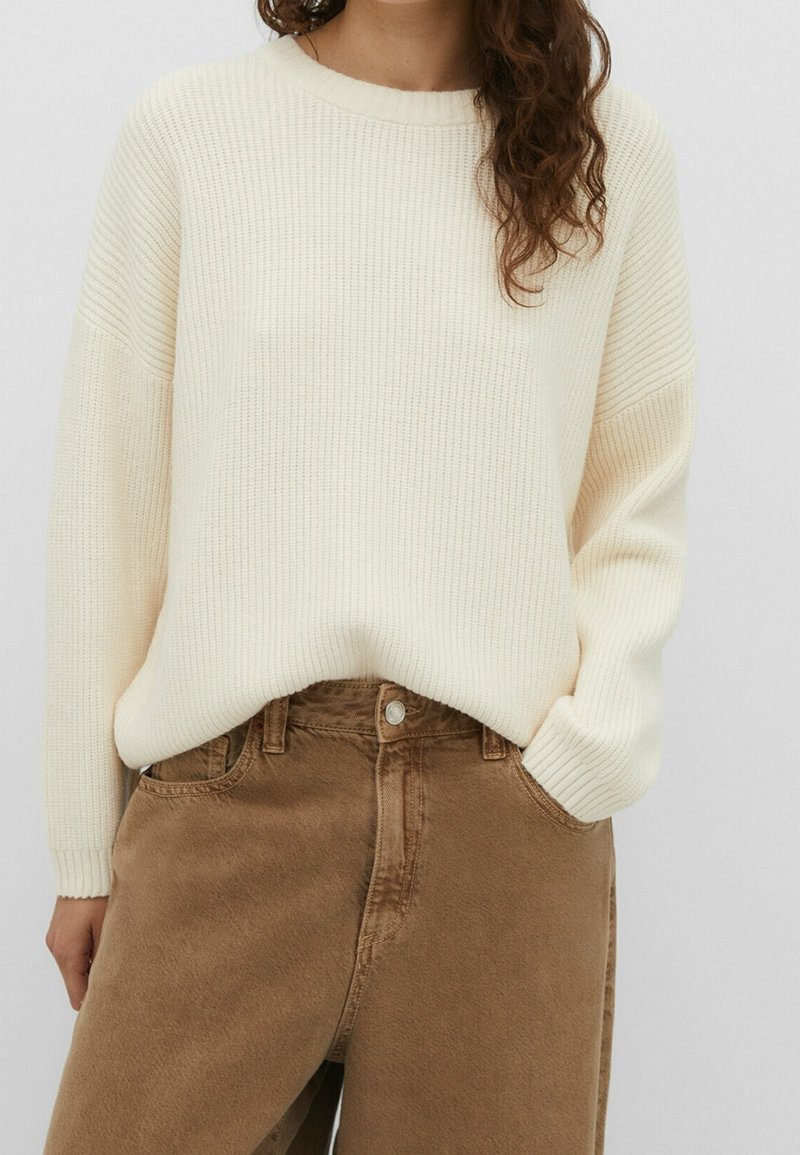 Personne portant un pull côtelé crème rentré dans un pantalon marron taille haute à jambes larges, avec une main dans la poche.