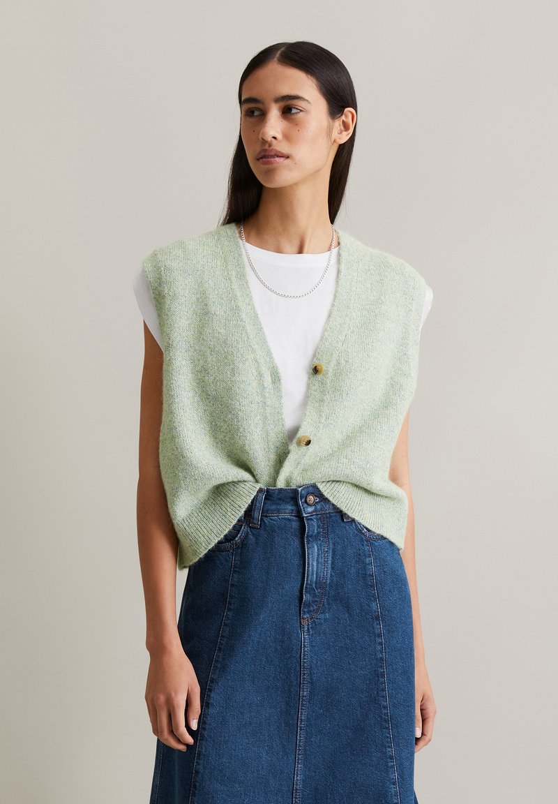 Gilet en tricot vert à manches courtes, avec un décolleté en V, trois boutons et une coupe ample, associé à une jupe en denim foncé.