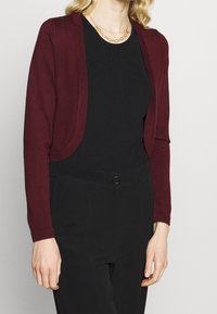 Burgundský cardigan s jemnou pletenou texturou, otevřeným designem a dlouhými rukávy, nošený přes černý žebrovaný top a černé kalhoty.