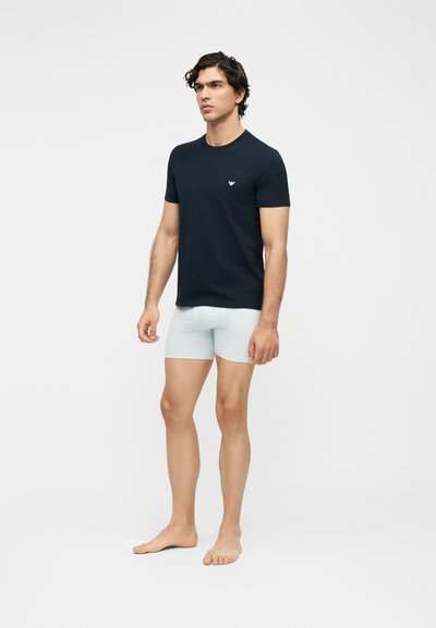 Άνδρας στέκεται ξυπόλυτος φορώντας μαύρο κοντομάνικο πουκάμισο με λογότυπο και ανοιχτό μπλε εφαρμοστά σορτς σε απλό ανοιχτό φόντο.