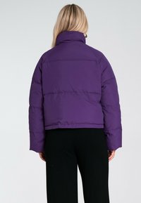 Fioletowa kurtka puchowa z wysokim kołnierzem, pikowanym wzorem i długimi rękawami. Tył ma skrócony krój i minimalne akcenty sprzętowe.