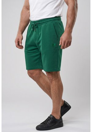 Mann trägt grüne Shorts mit Kordelzug und Seitentaschen sowie schwarze Sneakers vor hellem, einfachem Hintergrund.