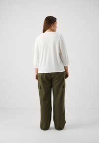 Balta mežģīņu blūze ar trīs ceturtdaļu piedurknēm, kas rotāta ar acu rakstiem. Saskaņota ar brīviem olīvu zaļas krāsas kargo biksēm.