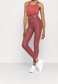 Débardeur rouge noué à la taille, associé à un legging rouge taille haute avec des fentes sur les côtés, rehaussé par des chaussures de sport blanches.