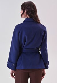 Giacca blu navy con tessuto textured, maniche ampie, vita arricciata e polsini con bottoni, caratterizzata da un colletto classico e una forma a peplo.