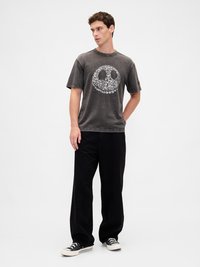 Juoda grafikos marškinėliai su taikos ženklu, pagaminti iš medvilnės, trumpomis rankovėmis ir laisvo silueto. Dėvimi su juodomis plačiomis kelnėmis ir sportbačiais.