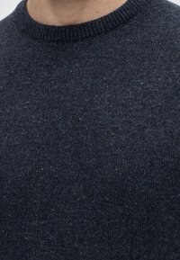 Gros plan d’un pull tricoté bleu foncé avec un col rond côtelé et de subtiles taches multicolores, porté par une personne.