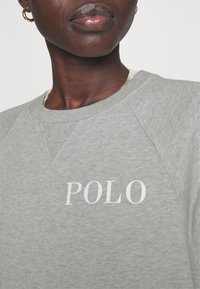 Šedá mikina s kulatým výstřihem a raglánovými rukávy. Na přední straně je vytištěno "POLO" bílou barvou. Materiál vypadá měkký a texturovaný.