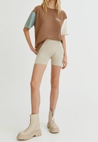 Donna in piedi con pantaloni ciclisti beige, stivali chunky beige con zip e una maglietta larga marrone con maniche multicolori.