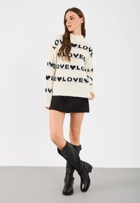 Gebreide trui met zwarte "LOVE" tekst en hartmotieven op een crèmekleurige achtergrond, gecombineerd met een zwarte rok en hoge zwarte laarzen.