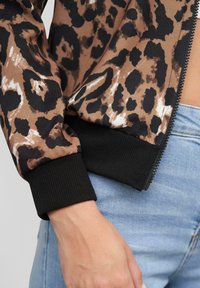 Giacca bomber a stampa leopardata marrone e nera con polsini ribattuti neri e cerniera. Presenta texture a contrasto tra i materiali.