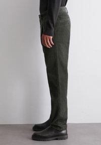 Tmavě zelené manšestrové kalhoty se rovný střih, texturovaný povrch a standardní kapsy, nošené s černými kotníkovými botami.