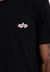 Sort bomulds t-shirt med et brystlogo, der har et hvidt stiliseret "A" og røde og hvide skrå striber. Kortsleeves, rund hals-design.