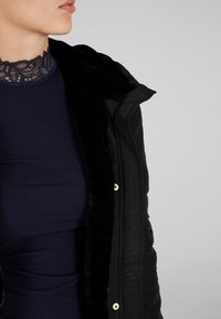 Černá prošívaná bunda s měkkým umělým kožíškovým límcem, doplněná tmavým žebrovaným pleteným topem s krajkovým výstřihem.