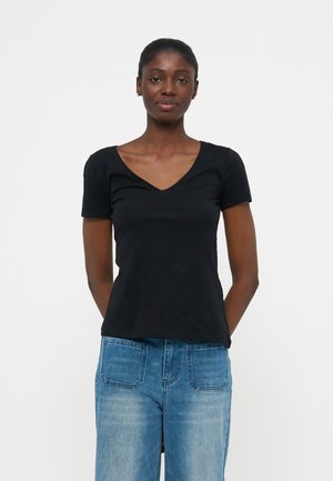 Γυναίκα στέκεται προς τα εμπρός φορώντας μαύρο κοντομάνικο μπλουζάκι με V λαιμόκοψη και μπλε τζιν, έχοντας τα χέρια πίσω από την πλάτη, σε απλό λευκό φόντο.