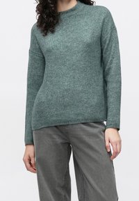 Groene geribbelde trui met een hoge hals, lange mouwen en een losse pasvorm, gemaakt van een zachte gebreide stof. Gecombineerd met grijze broek met wijde pijpen.