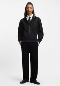 Person stående med hænderne i lommerne iført sort jakke med lynlås over hvid skjorte og sort slips, sorte bukser og sorte sko mod hvid baggrund.