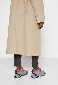 Personne portant un long manteau beige sur pantalon foncé et des chaussures de randonnée grises avec des lacets roses, debout devant un fond uni.