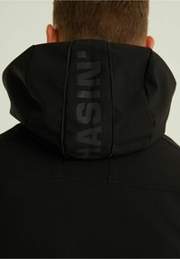 Homme portant une veste noire à capuche avec la capuche relevée, montrant un texte vertical discret "HASIN" au dos, sur un fond clair uni.