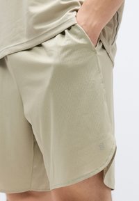 Μπεζ αθλητικό σορτς από ελαφρύ, διαπνέον ύφασμα με μικρές οπές. Διαθέτει πλαϊνή τσέπη και διακριτική λεπτομέρεια με λογότυπο.