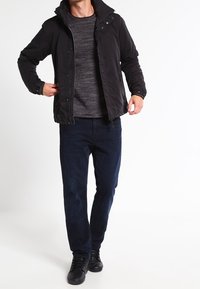 Veste noire légère avec capuche, chemise texturée grise, jean bleu foncé et baskets noires. La veste a des boutons-pression et une coupe décontractée.