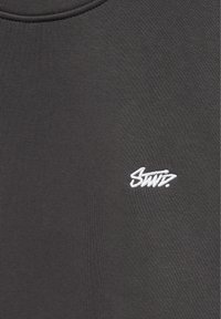 Gros plan sur un tissu gris foncé avec un petit texte stylisé brodé en blanc "Swiz" sur la zone de la poitrine d’un vêtement.