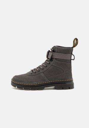 Bota alta de color gris oscuro con cordones en la parte delantera, correa ajustable, suela negra gruesa, costuras amarillas y lengüeta de tirador con marca en la parte trasera.