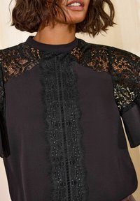 Sort top med blonder på skuldrene og et tekstureret blonderpanel foran. Korte ærmer og høj halsudskæring. Glat stoffinish.