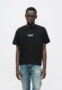 Černé bavlněné tričko s volným střihem, krátkými rukávy a bílým potiskem textu "CHALÉT SPORTING CLUB" na hrudi. Nošeno s světle modrými džínami.