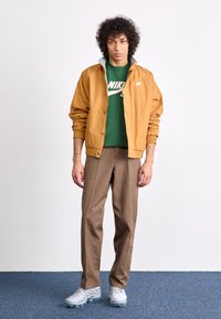 Tan windbreaker with elastic cuffs, layered over a green Nike T-shirt, paired with brown pants and grey athletic shoes with transparent soles.