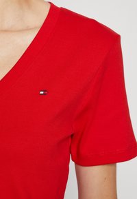 Röd V-ringad kortärmad skjorta som bärs på övre delen av kroppen med en liten rektangulär logotyp på vänster bröst.