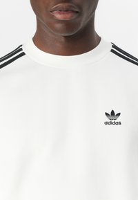 Bela majica s rundastim izrezom, crni trostruki detalji na ramenima i mali crni Adidas logo na prsima.