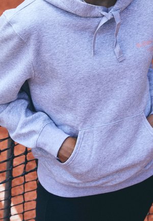 Person in grauem Hoodie mit den Händen in der vorderen Tasche, stehend neben einem schwarzen Netz über einem Sand-Tennisplatz.