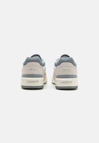 Sneakers Lacoste mají šedou a béžovou barevnou kombinaci, texturovaný síťovaný svršek, polstrovaný límec, retro design a embosované detaily loga.