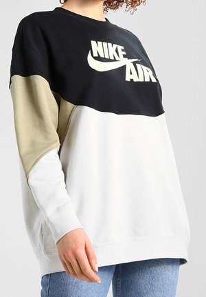 Personne portant un sweat-shirt Nike Air bicolore noir, beige et blanc avec un jean bleu clair, sur un fond gris uni.