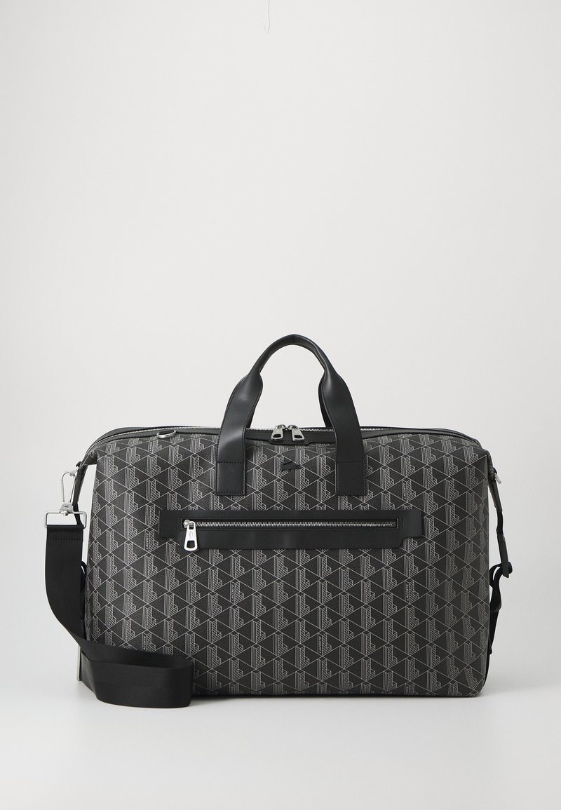 Sac de voyage noir à motifs avec doubles poignées, poche avant zippée et sangle d'épaule réglable sur un fond clair uni.