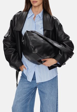 Kobieta ubrana w dużą czarną skórzaną kurtkę, jasnoniebieską koszulę w paski i dżinsy, trzymająca dużą czarną skórzaną torbę na ramię przewieszoną przez klatkę piersiową.