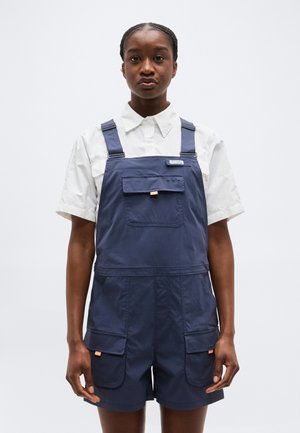 Γυναίκα που φοράει σκούρο μπλε κοντό φορμάκι με τσέπες στο μπροστινό μέρος και στους μηρούς, πάνω από ένα λευκό κοντομάνικο πουκάμισο με κουμπιά, στέκεται μπροστά σε λευκό φόντο.