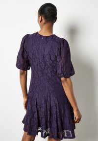 Fioletowa sukienka koronkowa z krótkimi bufkami i rozkloszowanym dołem. Detal wzoru koronkowego z kwiatami oraz ukrytym zamkiem błyskawicznym z tyłu.