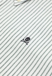 Polo a righe in cotone grigio chiaro e verde scuro, con un piccolo ricamo di teschio e ossa incrociate in nero sul lato sinistro.