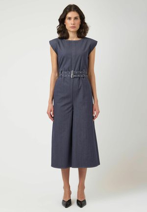 Kvinne med mørkt skulderlangt hår har på seg ermeløs marineblå dress med belte og svarte spisse hæler, stående mot ensfarget bakgrunn.