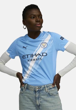 Light blue Manchester City jersey with short sleeves, featuring white stripes and black logos. Worn with light denim jeans.