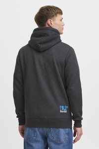 Sweat à capuche charbon avec une grande capuche ajustable, affichant un logo bleu et du texte dans le bas du dos, associé à un jean en denim bleu clair.