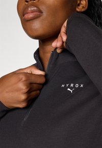 Giacca nera con zip, caratterizzata da un collo alto, un tessuto morbido e i loghi bianchi "HYROX" e Puma sul petto. Dettagli della zip ben visibili.